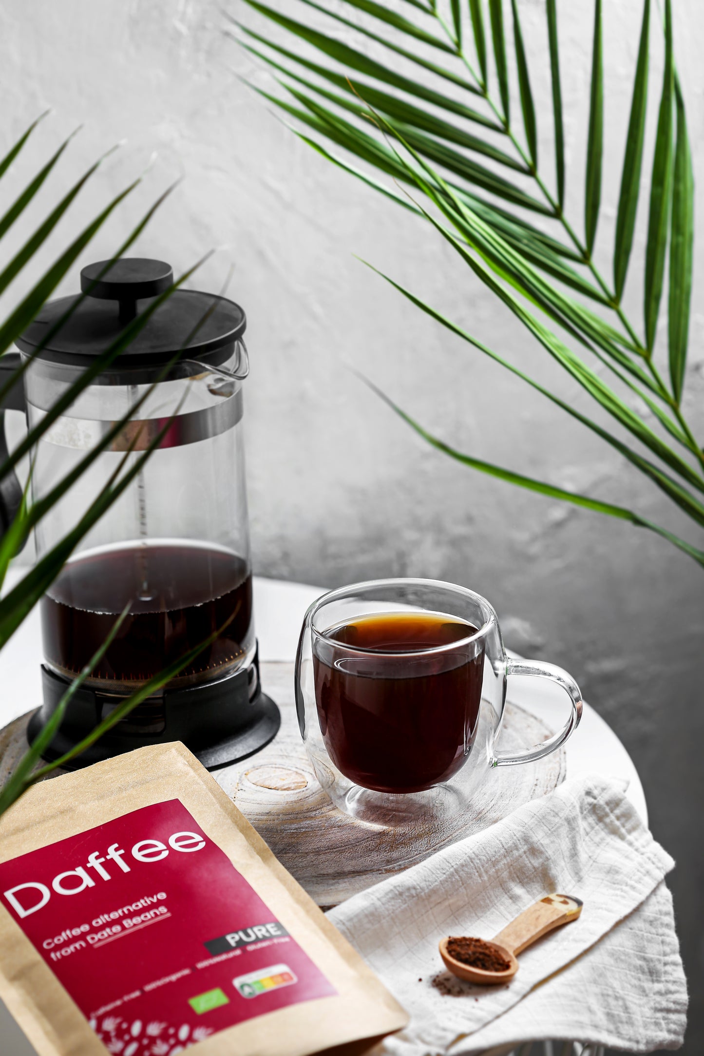 Daffee Pure alternatieve koffie van dadelbonen in glazen mok naast Franse pers, op linnen servet.